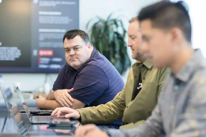 three men are sitting at a table with laptops in a meeting room.